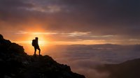 Silhouette of hiker standing on mountain peak at sunrise above clouds, adventure and exploration concept for travel and outdoor lifestyle content.