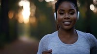 Young African American woman wearing wireless headphones and gray athletic top smiling outdoors with warm bokeh lights in background for fitness and wellness content.