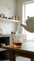 White ceramic teapot pouring hot tea into a glass cup on a wooden table in a cozy minimalist kitchen with neutral tones.