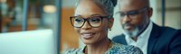 African American businesswoman with glasses and silver hair smiling while working at computer in modern office, mature male colleague in background.