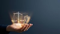 Hand holding glowing holographic scales of justice and security shield, symbolizing legal protection, cybersecurity law, and digital rights on dark background.