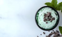Creamy mint ice cream topped with dark chocolate chunks and fresh mint leaves in black bowl on white background, overhead view with copy space.