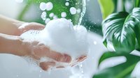 Hands holding white soap foam with water drops splashing against green tropical leaves background, fresh cleansing concept for skincare and hygiene products.