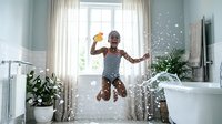 Young African American boy in striped shirt jumping joyfully in modern bathroom with water splashing around, expressing happiness and playful energy at home.
