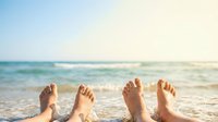 Two pairs of bare feet resting on a sunny sandy beach with ocean waves, evoking relaxation, summer vacation, and travel freedom.