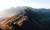 Majestic mountain peaks bathed in golden sunrise light, dramatic rocky cliffs and autumn forest slopes creating stunning natural landscape panorama.