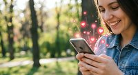 Young woman using smartphone with heart icons floating from screen in sunny park, social media engagement and digital communication concept.