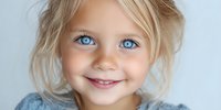 Portrait of young Caucasian girl with blonde hair and striking blue eyes smiling against soft blue background, conveying innocence and joy.