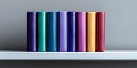 Colorful hardcover books arranged in row on white shelf against gray wall, creating minimalist composition with vibrant spines in purple, teal, blue and pink tones.