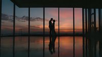 Romantic couple silhouette embracing by floor-to-ceiling windows at sunset with city skyline view. Modern office or apartment interior with reflection.
