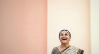 Mature woman with gray hair and South Asian ethnicity smiling upward against pink and cream wall background, expressing joy and positivity for lifestyle and wellness content.