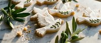 Elegant dove-shaped cookies with white icing and olive branch details arranged with fresh greenery on marble surface for peaceful celebrations.