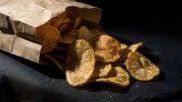 Crispy golden potato chips spilling from paper bag on dark slate surface with dramatic lighting for snack food advertising and packaging design.