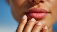 Close-up of woman's hand with manicured nails touching full lips against blue background, showcasing beauty and skincare concept.