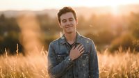 Young Caucasian man in denim shirt standing in golden wheat field at sunset, hand on chest, warm natural lighting, outdoor portrait for lifestyle and editorial use.
