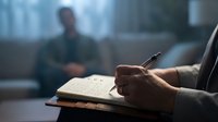 Professional businessman writing notes in journal during office meeting with colleague in blurred background for business planning and productivity.