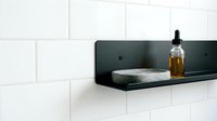 Floating black shelf with amber glass bottle and stone soap dish against white subway tile wall for bathroom storage and organization.