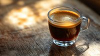 Fresh espresso shot with golden crema in clear glass cup on rustic wooden surface, natural sunlight casting soft shadows through window creates morning ambiance.