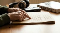 Elderly hands reading braille book with headphones and large print materials on wooden desk for visually impaired accessibility and learning.