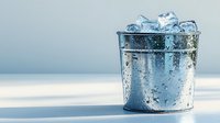 Ice cubes in vintage metal bucket with water drops and condensation on light blue background, minimalist composition with copy space for text.