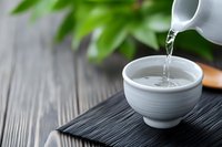 Pouring hot water from white ceramic teapot into traditional tea cup on wooden table with green plant background for relaxation and wellness ceremony.