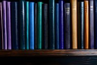 Row of colorful hardcover books arranged on wooden shelf, spines showing in shades of purple, turquoise, blue and brown creating gradient effect.