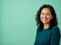 Woman with long dark curly hair and warm smile wearing teal sweater against mint green background, suitable for corporate portraits, business profiles, and professional headshots.