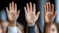 Three diverse raised hands showing palms against dark background, representing unity, voting, or participation in classroom or meeting setting.