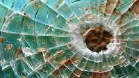 Shattered turquoise glass with circular impact hole, showing radiating cracks and jagged edges against rusty metal background.