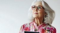 Senior woman with gray hair wearing glasses and floral shirt looking up thoughtfully against light background for lifestyle and aging concepts.