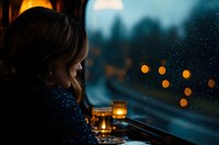 Young woman gazing through rainy window at city lights while sitting in cozy cafe with glowing candle, creating atmospheric moody evening scene.
