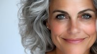 Confident mature woman with silver gray hair and bright smile showing natural aging beauty and self acceptance in close up portrait against light background.
