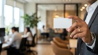 Businessman holding blank white business card in modern office environment with colleagues meeting in background, suitable for corporate branding and professional networking concepts.