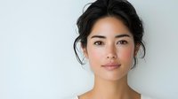 Young Asian woman with natural makeup and messy updo hairstyle looking at camera with confident smile against light background, beauty and skincare concept.