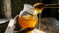 Honey dripping from wooden dipper into glass jar, golden liquid flowing, natural sweetener for cooking, baking, and wellness applications.