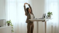 Professional businesswoman stretching arms above head at standing desk in bright modern office, taking wellness break during workday.