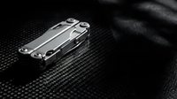 Stainless steel multi-tool with folded blades resting on textured black surface, suitable for camping, survival, and outdoor equipment design projects.
