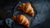 Fresh baked golden croissants on dark background with linen napkin, dramatic food photography with moody lighting and flaky pastry texture.