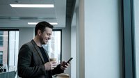 Professional Caucasian businessman in suit using smartphone while holding coffee cup in modern office building with large windows and city view.