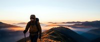 Hiker with backpack ascending mountain peak at sunrise, surrounded by misty valleys and golden light breaking through clouds overhead.