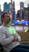 Middle-aged Caucasian man with beard sitting peacefully on bench with glowing green heart symbol on chest against blurred city skyline at dusk for wellness concepts.