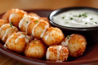 Crispy golden potato bites drizzled with creamy sauce and fresh herbs served on rustic brown plate with sour cream dipping sauce, close up food photography.