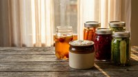 Homemade preserves and pickled vegetables in glass jars on rustic wooden table with warm sunlight streaming through kitchen window curtains.