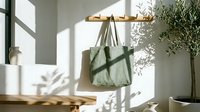 Green linen tote bag hanging on wooden wall rack in bright minimalist entryway with natural shadows and olive tree.