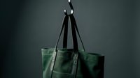 Dark green canvas tote bag with leather handles hanging on a metal hook against a moody gray background, showcasing minimalist style.