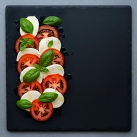 Fresh caprese salad with sliced tomatoes, mozzarella cheese and basil leaves arranged on dark slate board for Italian cuisine and healthy eating concepts.