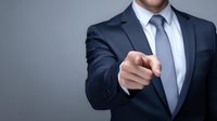 Confident businessman in navy suit pointing directly at viewer, conveying leadership and recruitment concept against neutral gray background.