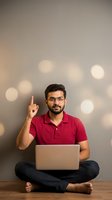 Young South Asian male professional wearing red polo shirt and glasses, sitting cross-legged with laptop, pointing upward with index finger against bokeh background. Suitable for tech, education, and business concepts.