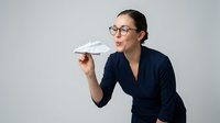 Young woman with dark hair, glasses, and olive complexion wearing a dark navy blazer, holding a paper airplane against a white background. Suitable for business, creativity, innovation, and startup concepts.