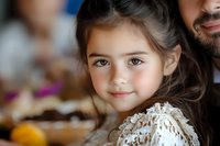 Young girl with long brown hair wearing lace dress smiles warmly at camera, close-up portrait showing bright eyes and natural expression against soft blurred background.